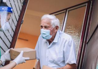 Young female volunteers in mask gives an elderly man boxes with food near his house